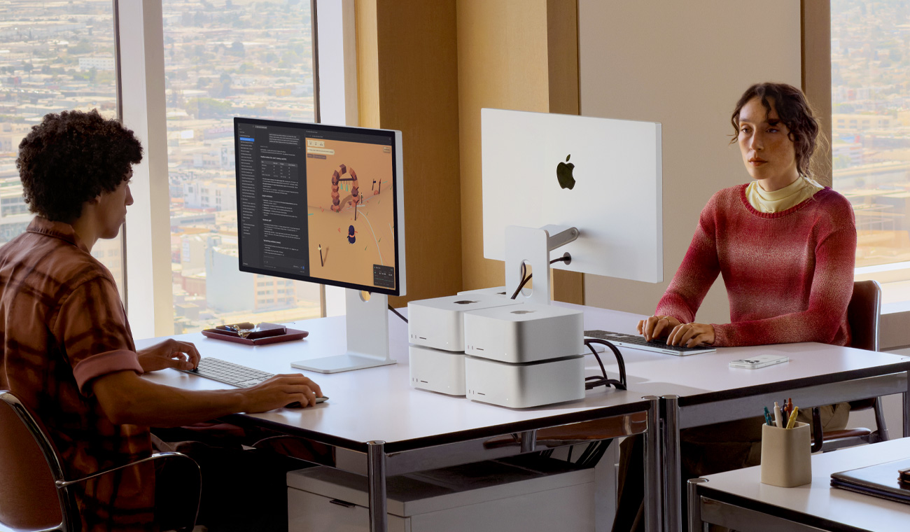 Two people sitting at two desks facing each other, four Mac Studios are on one desk in two stacks of two, two Studio Displays are on the desks, one screen shows various projects in-process in animation and editing apps, the other shows the back side of the display